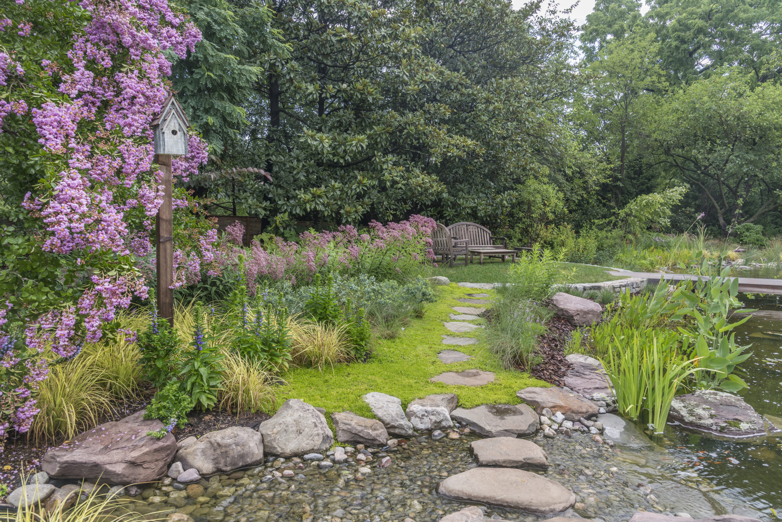 A tranquil garden with a stone path leading to a wooden bench, surrounded by lush greenery, blooming purple flowers, and a small pond with stepping stones and water plants. A wooden birdhouse on a post stands near the colorful blooms.