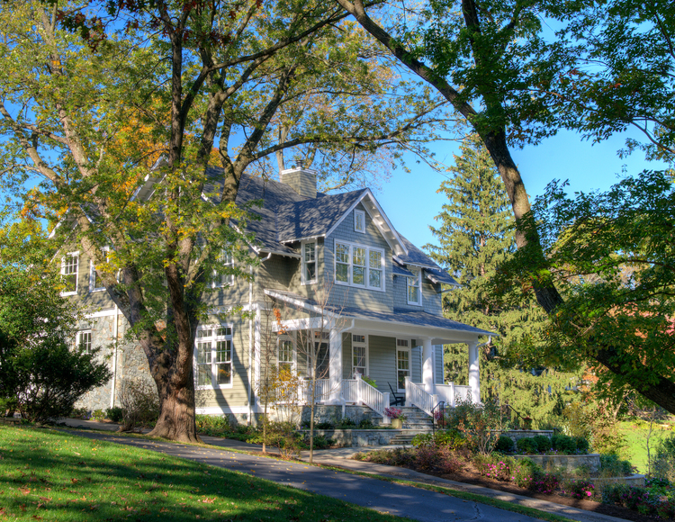 A two-story, pale green house with white trim and a covered porch, set against a backdrop of trees with autumn leaves, under a clear blue sky.