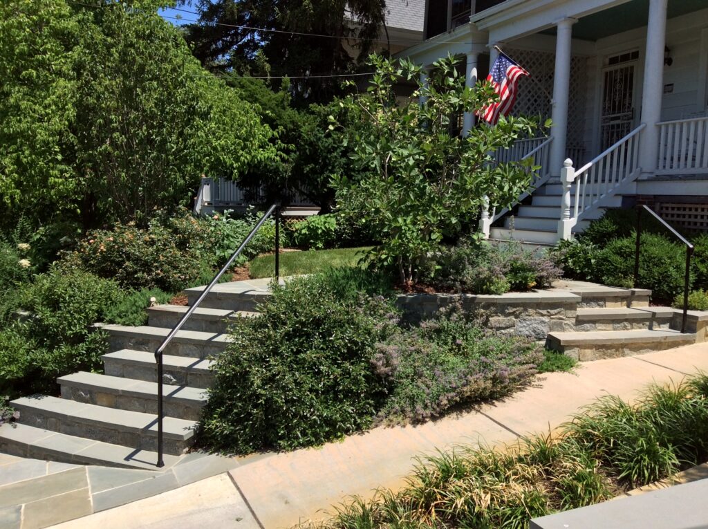 A landscaped front yard with a flagstone pathway leading to a white house with a front porch. An American flag hangs from the porch, and there are stone steps with metal railings surrounded by lush greenery and flowering shrubs.